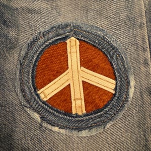May include: A denim patch featuring a peace symbol. The symbol is a white cross within a brown circle, surrounded by a blue denim border. The patch is sewn onto a light blue denim fabric.