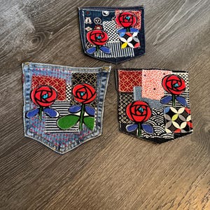 May include: Three denim pockets with patchwork designs and embroidered red roses. Each pocket features a unique combination of patterned fabrics, including checks, stripes, and floral prints. The roses have blue accents and green stems.