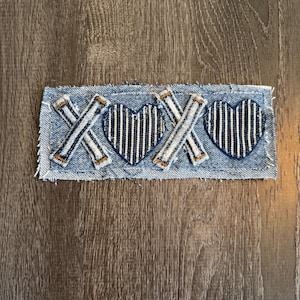 May include: A denim patch with two navy blue striped hearts and two "X" and "O" designs in a cross pattern. The patch has a frayed edge and is set against a wooden background.