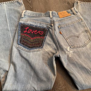 May include: Light blue denim jeans with a custom patch on the back pocket. The patch is embroidered with the word "Love" in red script and a heart. The jeans have a classic five-pocket design and a faded wash.