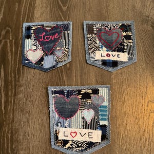May include: Three denim pocket patches with patchwork designs. Each patch features a heart design, with the word "Love" embroidered on two of them. The patches are made with various blue and white patterned fabrics.