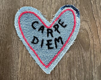 May include: A denim heart-shaped patch with the words "CARPE DIEM" embroidered in black. The heart is outlined in red stitching, and the edges are frayed. The patch is placed on a wooden surface.