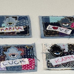Könnte beinhalten: Vier Denim-Patches mit gestickten Designs. Jeder Patch zeigt eine Hamsa-Hand, ein Auge und ein Wort: "Karma", "Love" und "Luck". Die Patches sind aus verschiedenen Stoffmustern und -farben gefertigt, darunter Blau, Weiß und Rot.