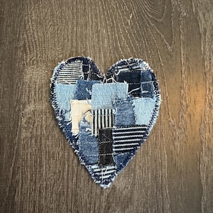 May include: A heart-shaped denim patch made from various shades of blue denim scraps. The patchwork design includes light and dark blue denim, with some pieces featuring striped patterns. The edges are frayed.