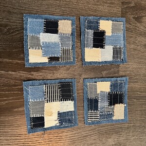 May include: Four square denim coasters with a patchwork design. Each coaster features a variety of denim shades, including light and dark blue, black, and cream, arranged in a geometric pattern. The edges are finished with a raw, frayed edge.