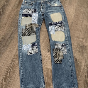May include: Blue denim jeans featuring a button fly closure and multiple rectangular fabric patches sewn onto the front legs. The patches consist of various patterns including dark blue paisley, black and white geometric prints, and striped fabric.