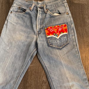 Könnte beinhalten: Hellblaue Jeans mit einem dekorativen Aufnäher auf der Gesäßtasche. Der Aufnäher zeigt ein rot-oranges Design mit einem Stierkopf und den darunter gestickten Worten "Hook 'Em". Die Jeans haben ein klassisches Fünf-Taschen-Design.