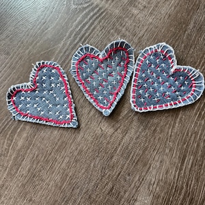 May include: Three denim heart-shaped patches with red stitching around the edges and white cross-stitch details. The patches are arranged on a wooden surface, showcasing a handmade craft aesthetic.
