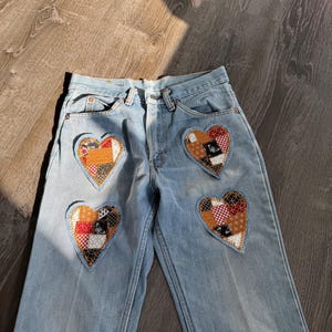 May include: Pale blue denim jeans featuring four heart-shaped patches. Each patch showcases a unique patchwork design with diverse colours and patterns, including red, orange, and brown. The jeans have a classic five-pocket design.
