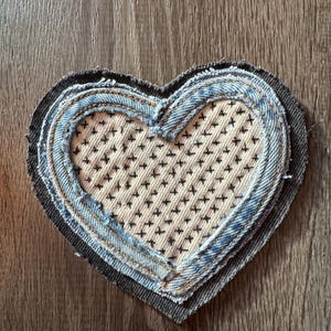 May include: A layered heart-shaped patch made from denim fabric. The heart features a light-colored center with black cross-stitch details, surrounded by denim layers in varying shades of blue and gray.