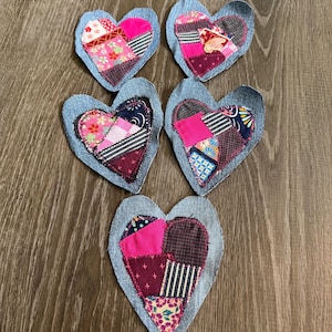 May include: Five heart-shaped fabric coasters. Each coaster features a patchwork design with denim blue borders. The patchwork includes pink, red, and patterned fabrics. The coasters are arranged on a wooden surface.