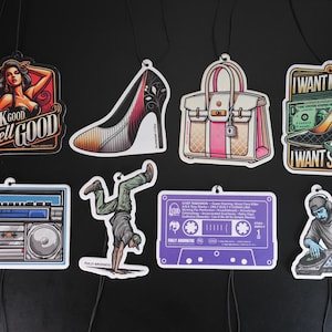 May include: A selection of air fresheners featuring diverse designs. The designs include a woman with the text "Look Good Smell Good", a high heel, a handbag, a boombox, a breakdancer, a cassette tape, and a DJ.