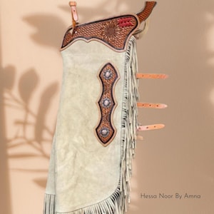 May include: Cream-colored leather chaps with intricate tooling and fringe detailing. The chaps feature a decorative sunflower design and three silver-toned embellishments. The text "Hessa Noor By Amna" is visible.