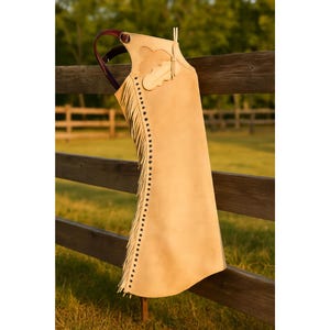 May include: A tan leather chaps with a decorative cloud and feather design. The chaps have fringe along the side and are hanging on a wooden fence. The chaps have a burgundy handle.
