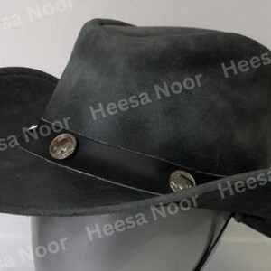 May include: A dark gray cowboy hat with a wide brim and a leather band. The hat features two decorative silver-colored buttons on the band. The hat is made of a soft, suede-like material.