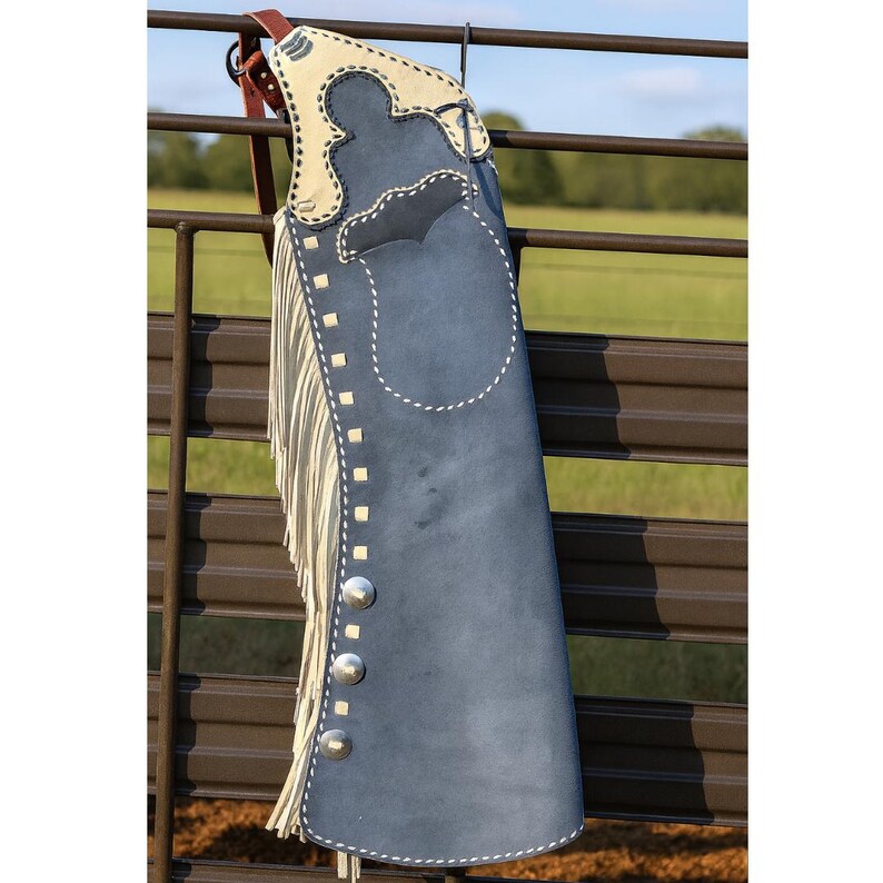 May include: A blue suede leather chaps with a cream-colored top section and fringe. The chaps have silver-colored button details and white stitching. The chaps are hanging on a brown metal fence.