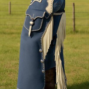 May include: Navy blue leather chaps with cream-colored fringe and accents. The chaps feature a pocket, decorative stitching, and silver-colored buttons. The person is wearing brown cowboy boots and blue jeans.