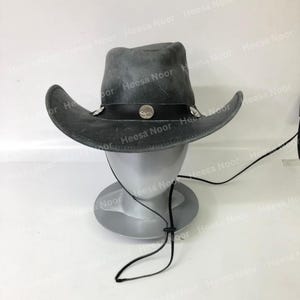 May include: A gray cowboy hat with a wide brim and a black leather band. The hat is decorated with silver-colored studs and a round emblem. It has a chin strap and is displayed on a mannequin head.