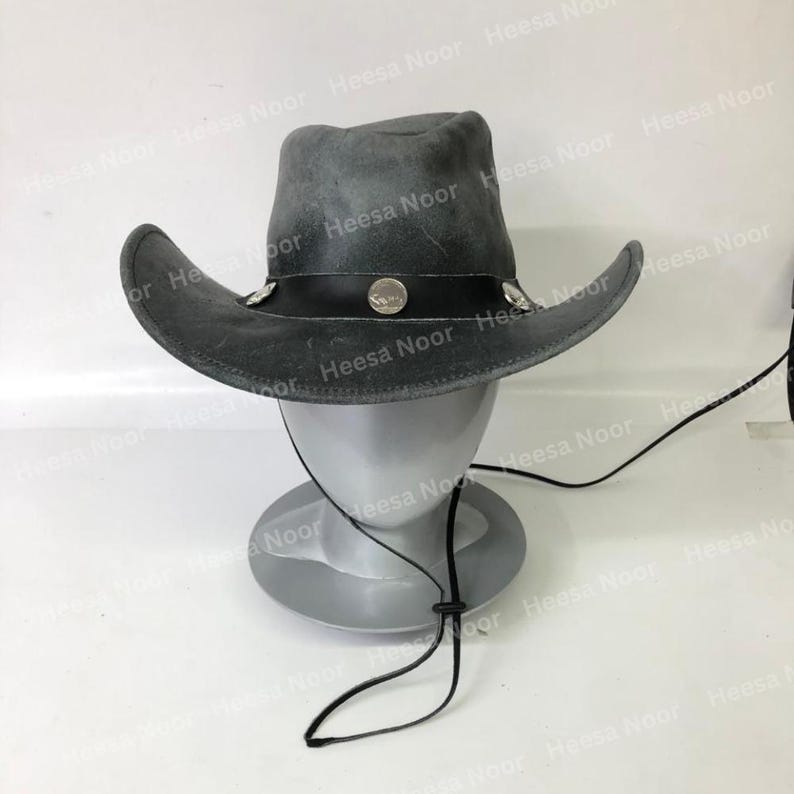 May include: A gray cowboy hat with a wide brim and a black leather band. The hat is decorated with silver-colored studs and a round emblem. It has a chin strap and is displayed on a mannequin head.