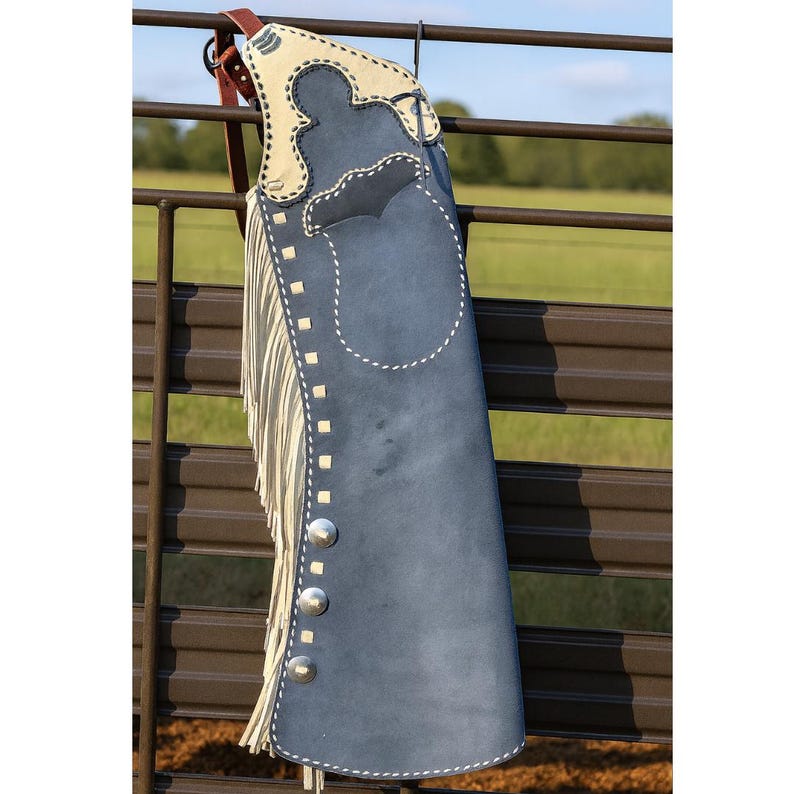 May include: A blue suede leather chaps with a cream-colored top section and fringe. The chaps have silver-colored button details and white stitching. The chaps are hanging on a brown metal fence.
