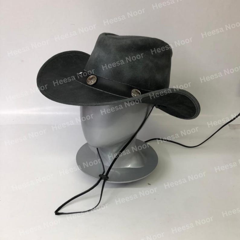 May include: A gray suede cowboy hat with a wide brim and a leather band. The hat features two decorative silver-colored buttons and a chin strap. The hat is displayed on a mannequin head.