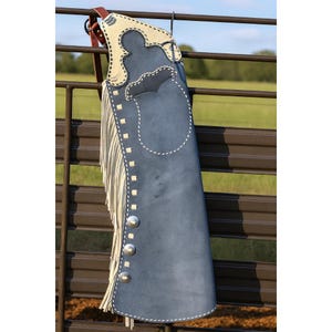 May include: A blue suede leather chaps with cream-colored accents and fringe. The chaps feature decorative stitching, silver-colored studs, and a leather strap. The chaps are hanging on a metal fence.