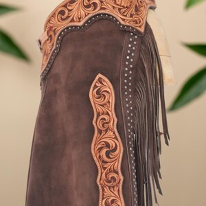 Op de afbeelding: Bruine leren chaps met ingewikkelde bloemen- en krulversieringen. De chaps hebben een decoratieve leren band aan de bovenkant, franjes langs de zijkant en zilverkleurige studs. De tekst "Hessa Noor by Amna" is zichtbaar.