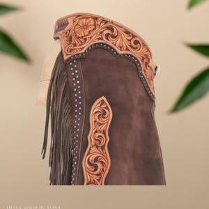 Op de afbeelding: Bruine leren chaps met ingewikkelde bloemen- en krulversieringen. De chaps hebben een franje langs de zijkant en decoratieve zilveren studs. De bovenste band is ook bewerkt met een bloemenmotief. De tekst "Hessa Noor by Amna" staat onderaan.