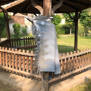 Puede incluir: Un par de polainas de cuero plateadas con flecos que cuelgan de la parte inferior. Las polainas están colgadas de un poste de madera.