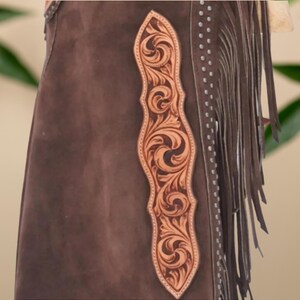 Op de afbeelding: Bruine leren chaps met een decoratief, lichtbruin leren paneel met bloemmotief. De chaps hebben franjes langs de zijkant en zijn versierd met zilverkleurige studs. De afbeelding bevat de tekst "Hessa Noor by Amna".