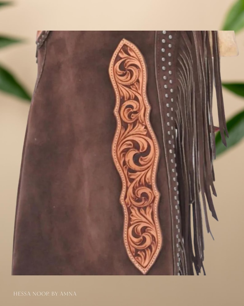 Op de afbeelding: Bruine leren chaps met een decoratief, lichtbruin leren paneel met bloemmotief. De chaps hebben franjes langs de zijkant en zijn versierd met zilverkleurige studs. De tekst "Hessa Noor by Amna" is zichtbaar onderaan.