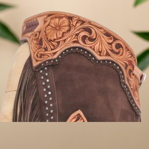 Op de afbeelding: Close-up van een bruine leren rijbroek met ingewikkelde bloemen- en krulversieringen. De chaps hebben een decoratieve bovenband met een bloemenmotief en zilveren studs. De tekst "HESSA NOOR BY AMNA" is zichtbaar aan de onderkant.