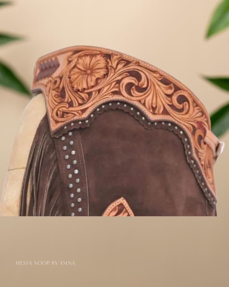 Op de afbeelding: Close-up van een bruine leren rijbroek met ingewikkelde bloemen- en krulversieringen. De chaps hebben een decoratieve bovenband met een bloemenmotief en zilveren studs. De tekst "HESSA NOOR BY AMNA" is zichtbaar aan de onderkant.