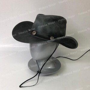 May include: A dark gray cowboy hat with a wide brim and a leather band. The hat is decorated with two silver-colored studs. A black chin strap is attached. The hat is displayed on a mannequin head.