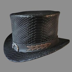 Puede incluir: Un sombrero de copa negro con un patrón texturizado de piel de serpiente en la corona y el ala. Una banda negra lisa rodea la base, decorada con un emblema de metal plateado de dos pistolas cruzadas y un detalle de cordones de cuero marrón.