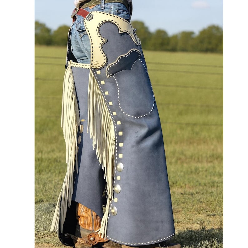 May include: Blue suede cowboy chaps with cream-colored accents and fringe. The chaps feature decorative stitching and silver-colored snaps. The chaps are worn over blue jeans and brown cowboy boots, suggesting a Western theme.