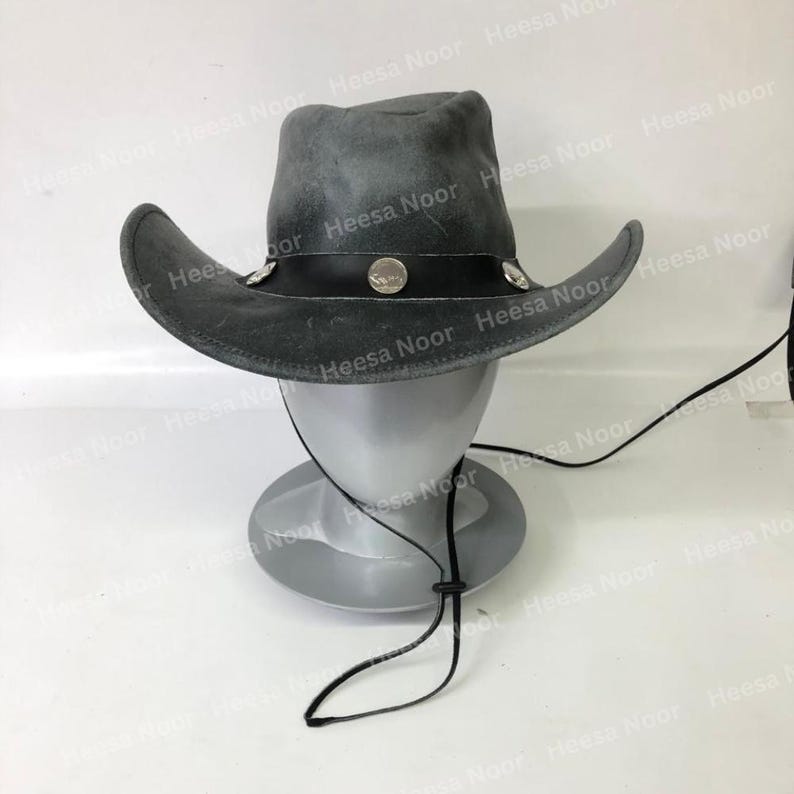May include: A gray cowboy hat with a wide brim and a black band. The hat is decorated with silver-colored studs and a round emblem. A black chin strap is attached. The hat is displayed on a mannequin head.