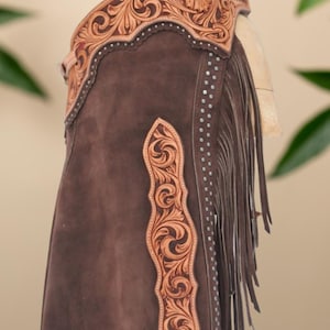 Op de afbeelding: Bruine leren chaps met ingewikkelde bloemen- en krulversieringen. De chaps hebben een decoratieve leren band aan de bovenkant, franjes langs de zijkant en zilverkleurige studs. De tekst "Hessa Noor by Amna" is zichtbaar.