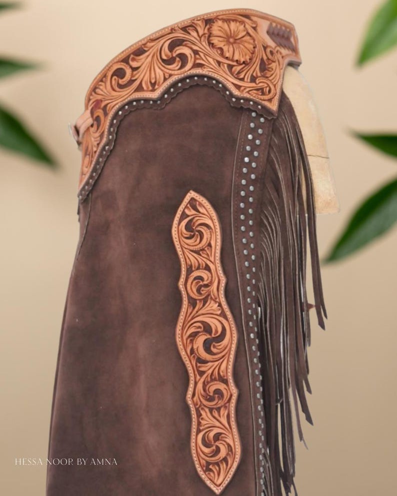 Op de afbeelding: Bruine leren chaps met ingewikkelde bloemen- en krulversieringen. De chaps hebben een decoratieve leren band aan de bovenkant, franjes langs de zijkant en zilverkleurige studs. De tekst "Hessa Noor by Amna" is zichtbaar.