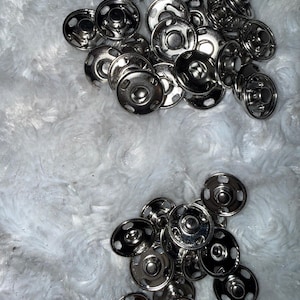 May include: A collection of shiny, silver-colored snap fasteners. The snaps are round with a central stud and multiple small holes around the edge. They are scattered on a fluffy, white surface.