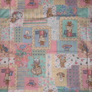 May include: A cotton fabric featuring a patchwork design with repeating illustrations of rabbits in various poses, lit candles, and floral patterns in soft pastel shades of pink, blue, yellow, and green, arranged in a grid of rectangular blocks.