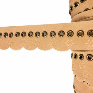 May include: A roll of beige faux suede trim featuring a scalloped edge with embossed flower patterns and a row of evenly spaced antique brass metal grommets running along the top length of the material.