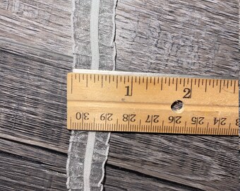 May include: White ruffled trim with a silver shimmer, measured against a wooden ruler. The trim is approximately 1 inch wide.