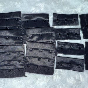 May include: A collection of black bra hook and eye closures in various sizes. The closures are made of a shiny, woven fabric and feature multiple hook and eye sets. The image shows a variety of bra making supplies.