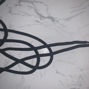 May include: Black shoelaces, intricately knotted, against a white and gray marbled background. The laces are thick and appear to be made of a durable, woven material. The tips of the laces are visible, suggesting they are ready to be tied.