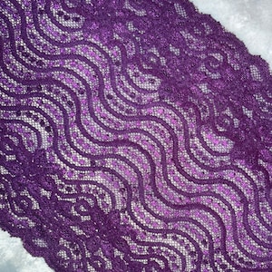 May include: Close-up of a length of purple lace with a wavy pattern. The lace has a delicate, openwork design with a repeating motif of swirls and dots. The color is a deep, rich purple with a subtle sheen, and the lace is set against a white background.