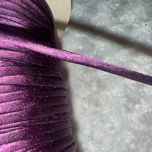 May include: A spool of purple satin cord with a shiny finish. The cord is wound tightly and is ready to be used for crafting or other projects.