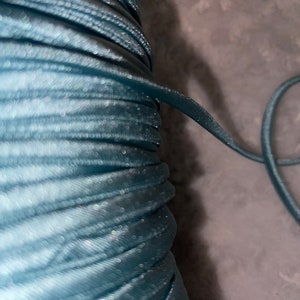 May include: A close-up of a spool of teal blue elastic cord. The cord is wound tightly and has a slight sheen.