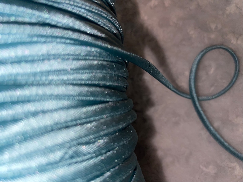 May include: A close-up of a spool of teal blue elastic cord. The cord is wound tightly and has a slight sheen.