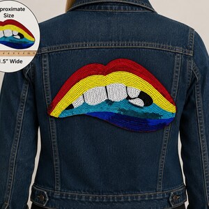 May include: A dark blue denim jacket featuring a large, sequined patch on the back in the shape of lips. The lips are colored in horizontal stripes of red, yellow, light blue, and dark blue, with white teeth. A ruler shows it is 11.5 inches wide.